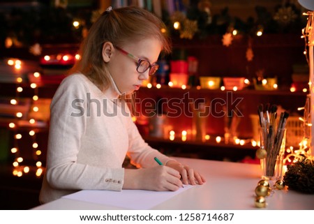 Blonde child girl with big pink ans black glasses writing letter to the Santa Claus or drawing something on the background with warm yellow bokeh. Christmas and New Year theme.