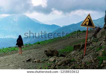 Young woman with backback hiking through the way in high mountains under yellow warning sign