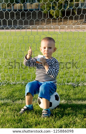 Small boy sitting on a soccer ball in a goalpost giving a thumbs up sign of approval with his hand