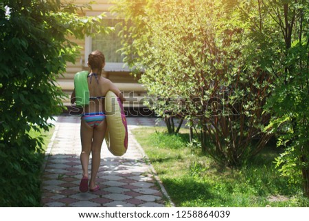 Teenager girl in a bathing suit with an inflatable pool ring walks along a stone path among trees and grass.