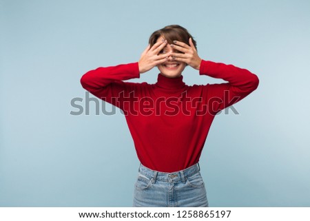 Beautiful smiling girl with dark short hair in red sweater and jeans happily covering eyes with hands over blue background 