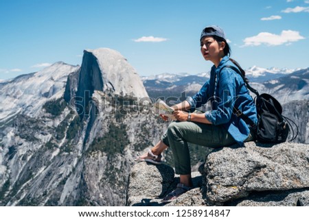 cool female backpacker sitting on the big rock on top of mountain in yosemite national park. young asian hiker holding paper map checking the destination in hiking travel. woman backpacker enjoy view