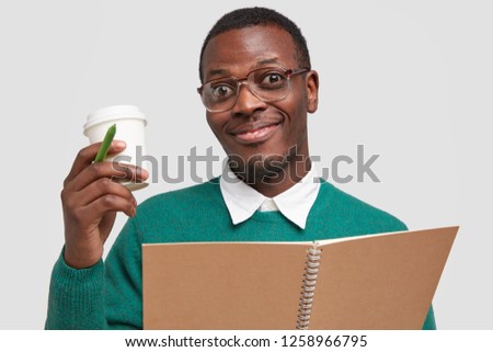 Horizontal shot of pleased dark skinned young man with stubble, wears square spectacles, holds takeaway coffee, pen and notebook, pleased with good working result, isolated over white background