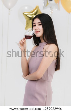 Attractive young asian woman in pink dress, drinking wine and happy at fun party, portrait on white background with colorful festive balloons.