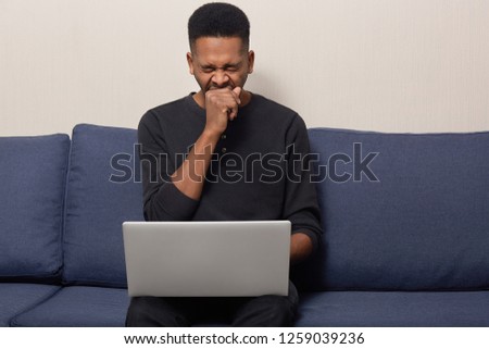 Photo of tired dark skinned feels fatigue of work on laptop computer, yawns and wants to sleep, dressed in black sweater, sits at sofa, prepares business project for tomorrow, being overworked