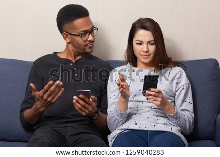 Indoor shot of mixed race woman and man search web pages on mobile phones, update software, have puzzled facial expression as have some problems with internet connection. Technology concept.