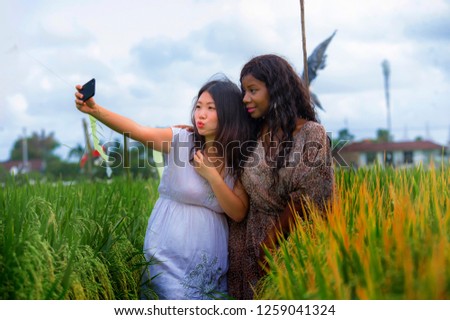mixed ethnicity Asian Chinese girl and black African American woman taking girlfriends selfie with mobile phone together isolated at rice field enjoying exotic tropical holidays in traditional village