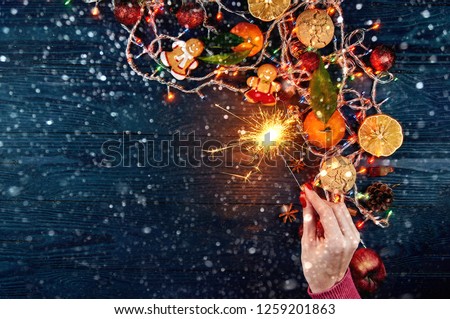 New Year's layout on a black wooden background with free space for text. A beautiful female hand in a pink winter sweater keeps a sparkler fire over the Christmas accessories.