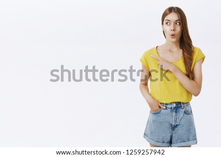 Wow looks cool. Amused and excited attractive caucasian female in yellow t-shirt folding lips from amazement and surprise gazing and pointing left with curious and interested look over white wall