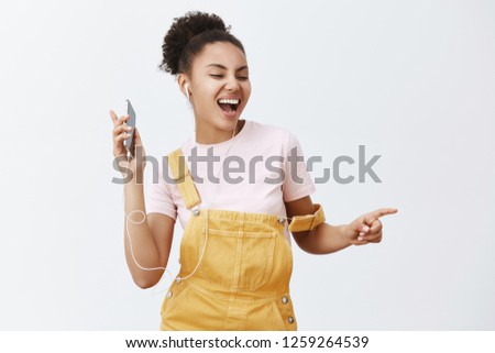 Wanna dance with somebody. Portrait of joyful happy attractive African American woman in yellow stylish dungarees, moving in rhythm of music, listening songs in earphones, holding cellphone