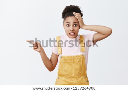 Yikes, what is that. Portrait of shocked worried and confused attractive african-american female with curly hair in fashionable yellow overalls, holding hand on head, pointing left with index finger Royalty-Free Stock Photo #1259264908