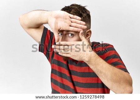 Funny stylish young man wearing red and black striped t-shirt posing indoors with hands at his face forming rectangular frame with only eye being seen, staring at camera as if taking picture