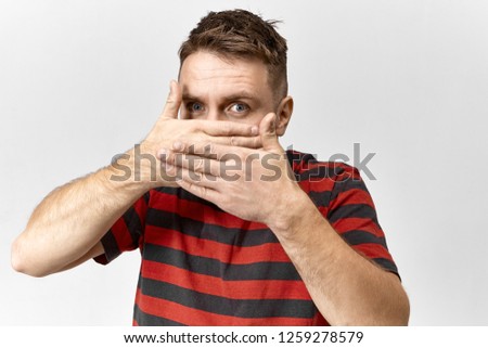 Secrecy and fear concept. Isolated view of mysterious young man in striped t-shirt covering his mouth, forbidden to speak, not allowed to reveal confidential information or secret, being intimidated Royalty-Free Stock Photo #1259278579