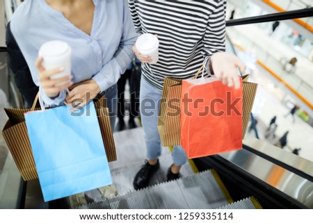 Girls with drinks and paperbags chatting while moving upstairs on escalator inside modern trade center during shopping