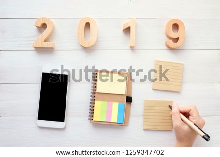 2019 wood letters, hand holding pen over blank notebook paper, white smart phone with blank screen on white wood background, 2019 new year mock up, template with copy space for text, top view
