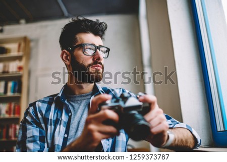 Thoughtful bearded hipster guy in eyeglasses looking out of window while holding vintage camera and making editing of photos on device.Pondering creative photographer dressed in casual shirt