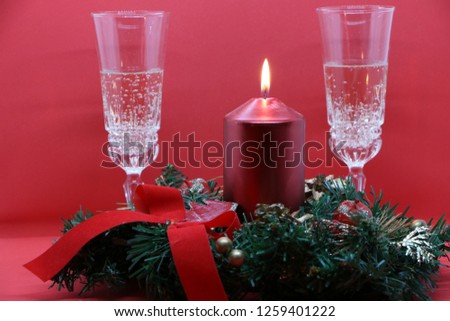 Two glasses of champagne, a burning red candle, a wreath on a red background.