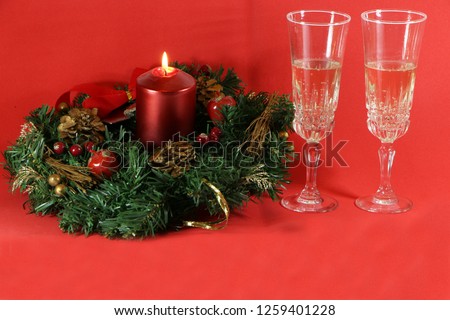 Two glasses of champagne, a burning red candle, a wreath on a red background.