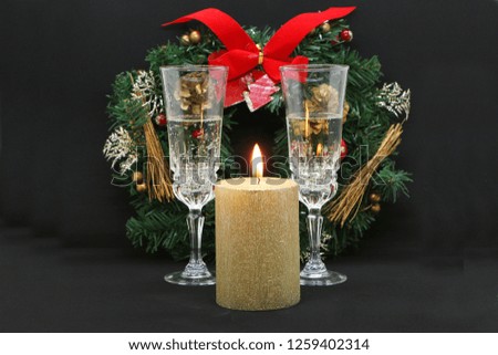 Two glasses of champagne, a burning golden candle, a wreath on a black background.