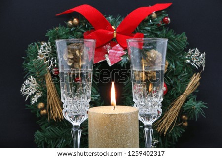 Two glasses of champagne, a burning golden candle, a wreath on a black background.