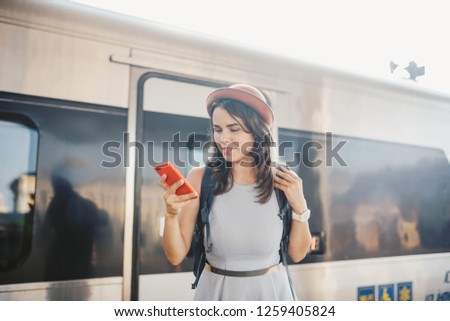 Theme transportation and travel. Portrait young caucasian woman with toothy smile standing train station train background with backpack using technology, smart phone hands in dress and hat summer.