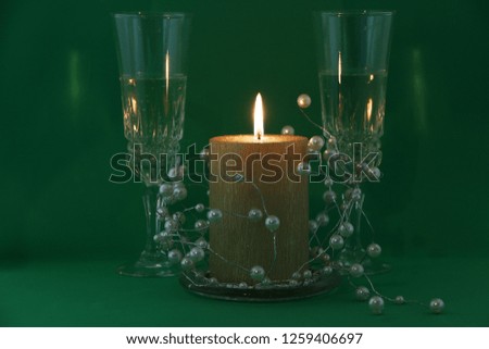 Two glasses of champagne, a burning golden candle, a wreath on a green background.