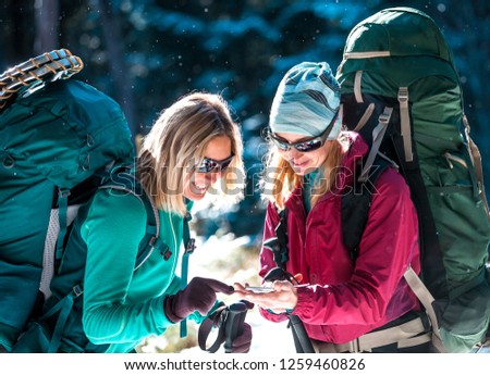 Two tourists look at the phone. Two friends travel together. Women with backpacks in the winter mountains. Winter hike. Walk through the snowy forest. Mobile communication and navigation.