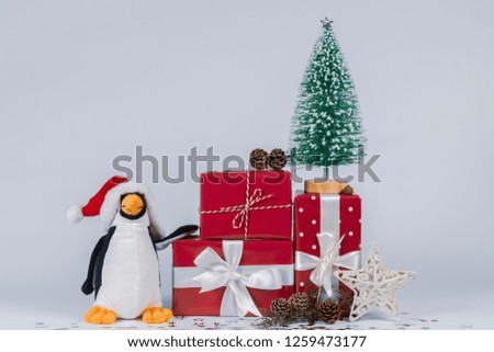 A funny penguin with Santa Claus hat stands near a little Christmas tree on gift packs with snow and shining little stars, pine cones