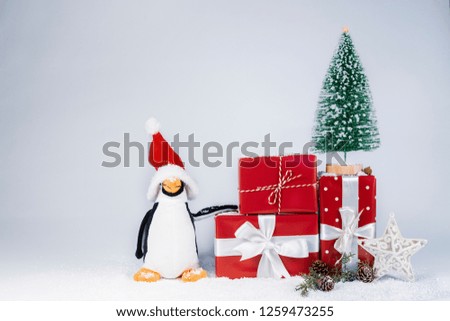A funny penguin with Santa Claus hat stands near a little Christmas tree on gift packs with snow and shining little stars, pine cones