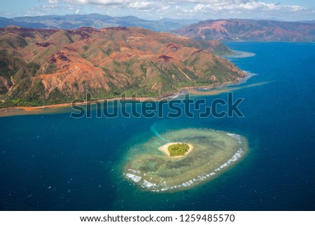 A small atoll islet with heart shaped coral reef off the east coast of Grande Terre island of New Caledonia, French overseas collectivity. Red green mountains hills full of nickel ore near Nakéty. Royalty-Free Stock Photo #1259485570
