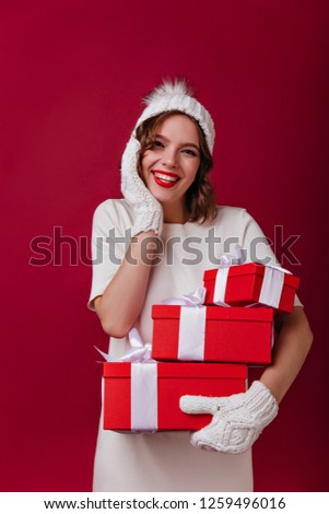Happy female model in knitted accessories laughing during winter photoshoot. Enchanting girl in hat holding christmas gifts and smiling.
