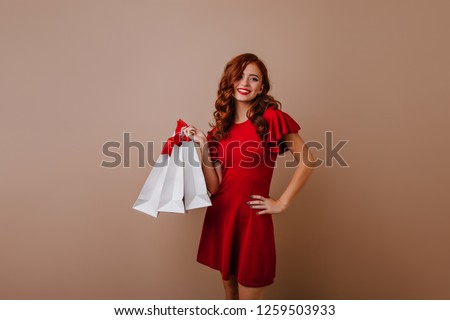 Shapely red-haired girl posing after shopping. Studio shot of female shopaholic wears red dress.