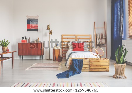 Wooden bed with white bedding and red pillow in spacious bedroom with striped carpet and retro furniture, real photo