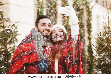Christmas selfie: young happy smiling couple takes photo on smartphone, posing on street. Models bundled up tartan blanket, holding candy canes. Snowfall. Empty, copy space for text