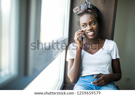 Cheerful afro american woman talking phone and sitting on windowsill