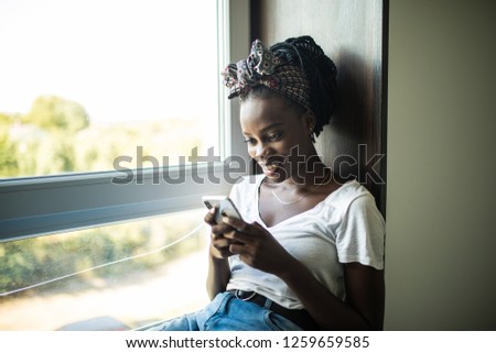 Smiling afro american woman sitting on the windowsill and writing message on phone at home