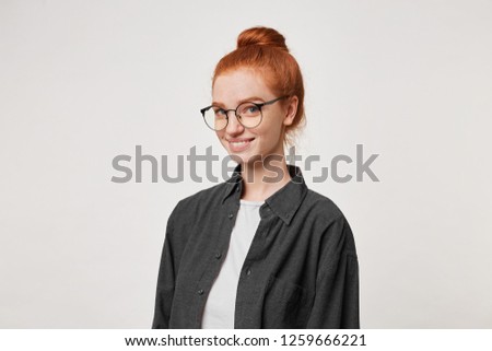 A young red-haired girl stands in a half-turn looking at the camera through glasses.Cheerful young woman dressed in a black man's shirt is smiling affably isolated on a white background.
