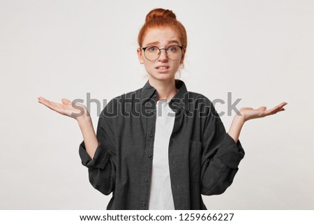 Studio photo of a girl with red hair wearing glasses dressed in a man's black shirt depicts helplessness hopelessness holding hands to the sides with her palms upwards. Confused scared apologizes.