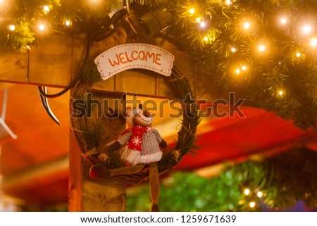 funny elk figure with pullover and red scarf sits in a wreath made of birch branches and sign "welcome", which hangs on a stall at the christmas market surrounded by christmas lights and fir