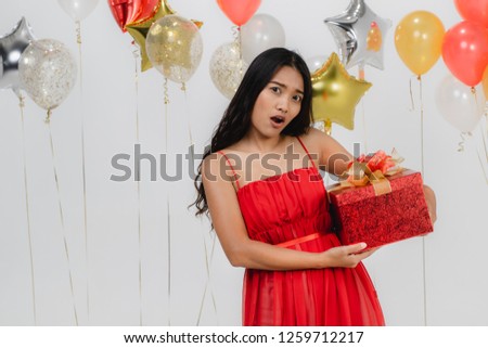 Attractive young asian woman wears red dress excited with her gift, happy at fun party, portrait shot white background with festive balloon.