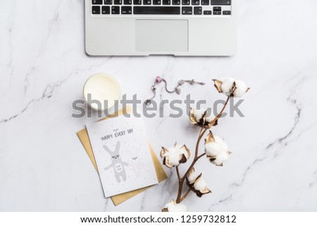 Flatlay items: laptop, cotton flower, card, candle lying on white marble background. Top view