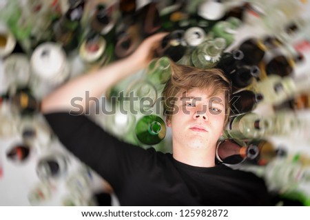 Young man with short blond hair lying on the floor and is surrounded by many empty beer and liquor bottles, upper perspective. Royalty-Free Stock Photo #125982872