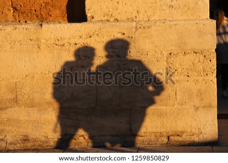 Outdoor view of two silhouettes of men walking in an urban street. Shadows of 2 male pedestrians projected on an ancient textured stone wall. Human shapes drawn on a brown lighted masonry surface.