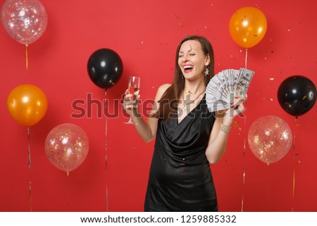 Laughing woman in black dress celebrating, holding bundle lots of dollars, cash money, glass of champagne on bright red background air balloons. Happy New Year, birthday mockup holiday party concept