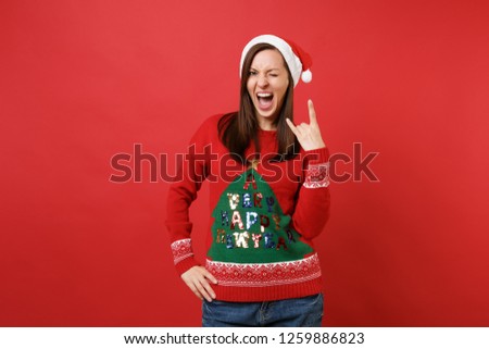 Cheerful Santa girl screaming, showing horns gesture, depicting heavy metal rock sign, blinking isolated on red background. Happy New Year 2019 celebration holiday party concept. Mock up copy space