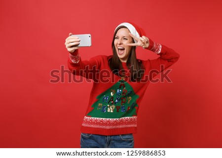 Funny Santa girl with opened mouth doing taking selfie shot on mobile phone, showing victory sign isolated on red background. Happy New Year 2019 celebration holiday party concept. Mock up copy space