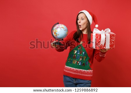 Shocked young Santa girl keeping mouth wide open, holding world globe, red box with gift, present isolated on red background. Happy New Year 2019 celebration holiday party concept. Mock up copy space