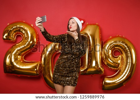 Merry Santa girl in shiny glitter dress, Christmas hat doing selfie shot isolated on bright red wall background, golden numbers air balloons studio portrait. Happy New Year 2019 holiday party concept
