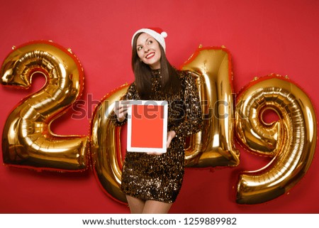 Merry Santa girl in shiny glitter dress, Christmas hat hold tablet pc isolated on bright red wall background, golden numbers air balloons studio portrait. Happy New Year 2019 holiday party concept