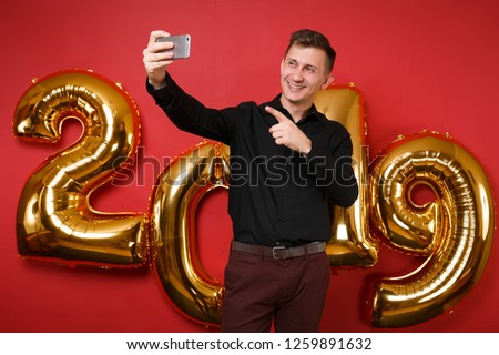 Merry fun young man in black shirt celebrating holiday party hold cellphone isolated on bright red wall background, golden numbers air balloons studio portrait. Happy New Year 2019 Christmas concept